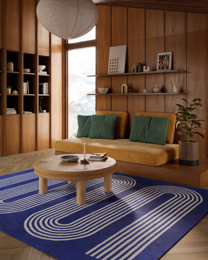 Wood-paneled living room with a mustard-yellow sofa, green cushions, round wood coffee table, blue concentric rug (5' x 8'), shelves and a plant.