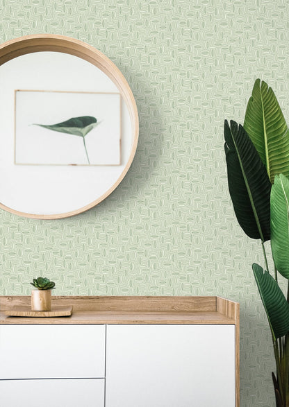 Geometric green matte wallpaper backdrop behind a round wooden mirror and a light wood console with white drawers; small succulent and tall leafy plant.