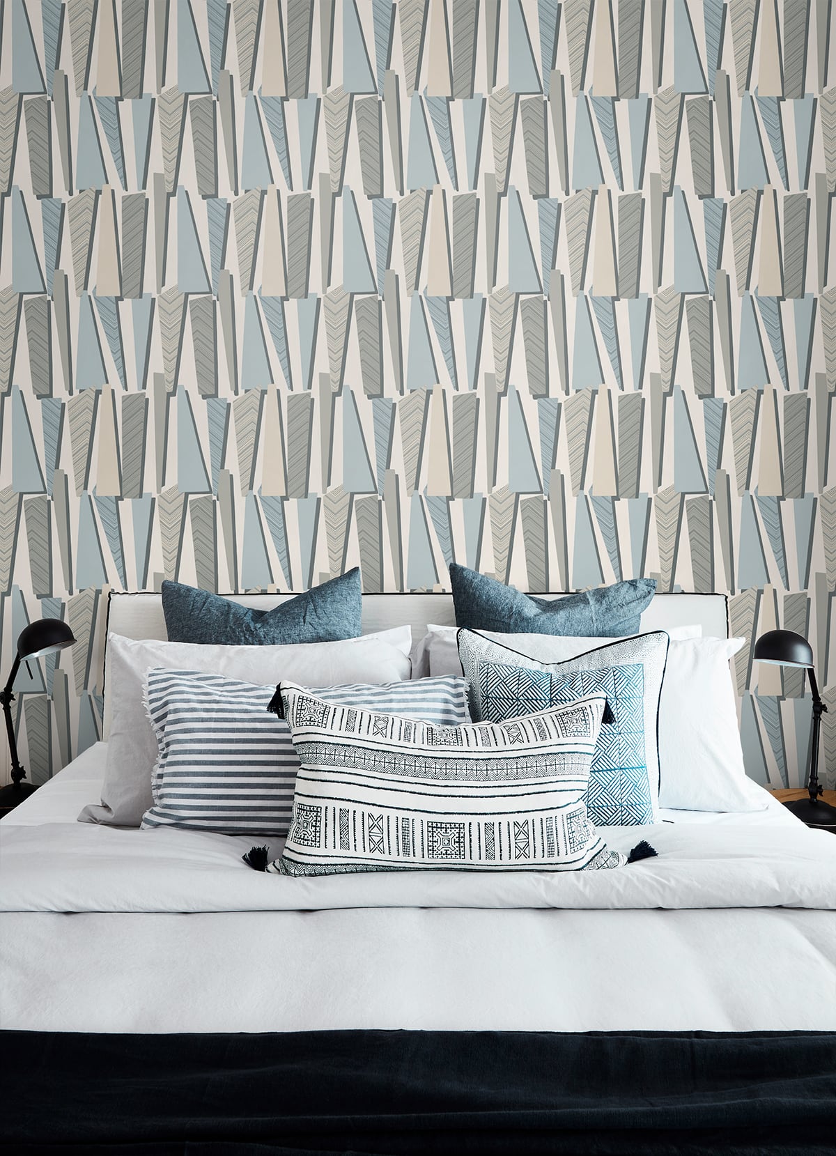 Queen bed with white duvet, blue pillows, striped and geometric tassel lumbar cushion, against blue-gray chevron wallpaper.