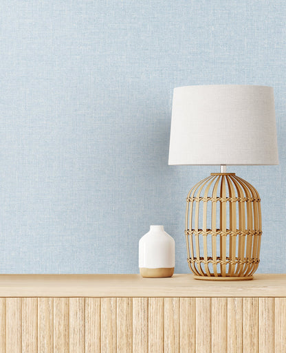 Soft blue matte linen textile product shown in a room setting with a boho rattan table lamp on a light wood sideboard, a small white ceramic vase, and a blue textured wall.
