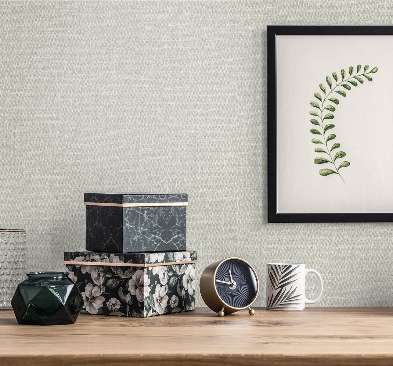 Flat lay with soft grey linen background featuring a stack of black floral and marble boxes, brass clock, faceted dark vase, striped mug, and a framed botanical print on a gray wall.