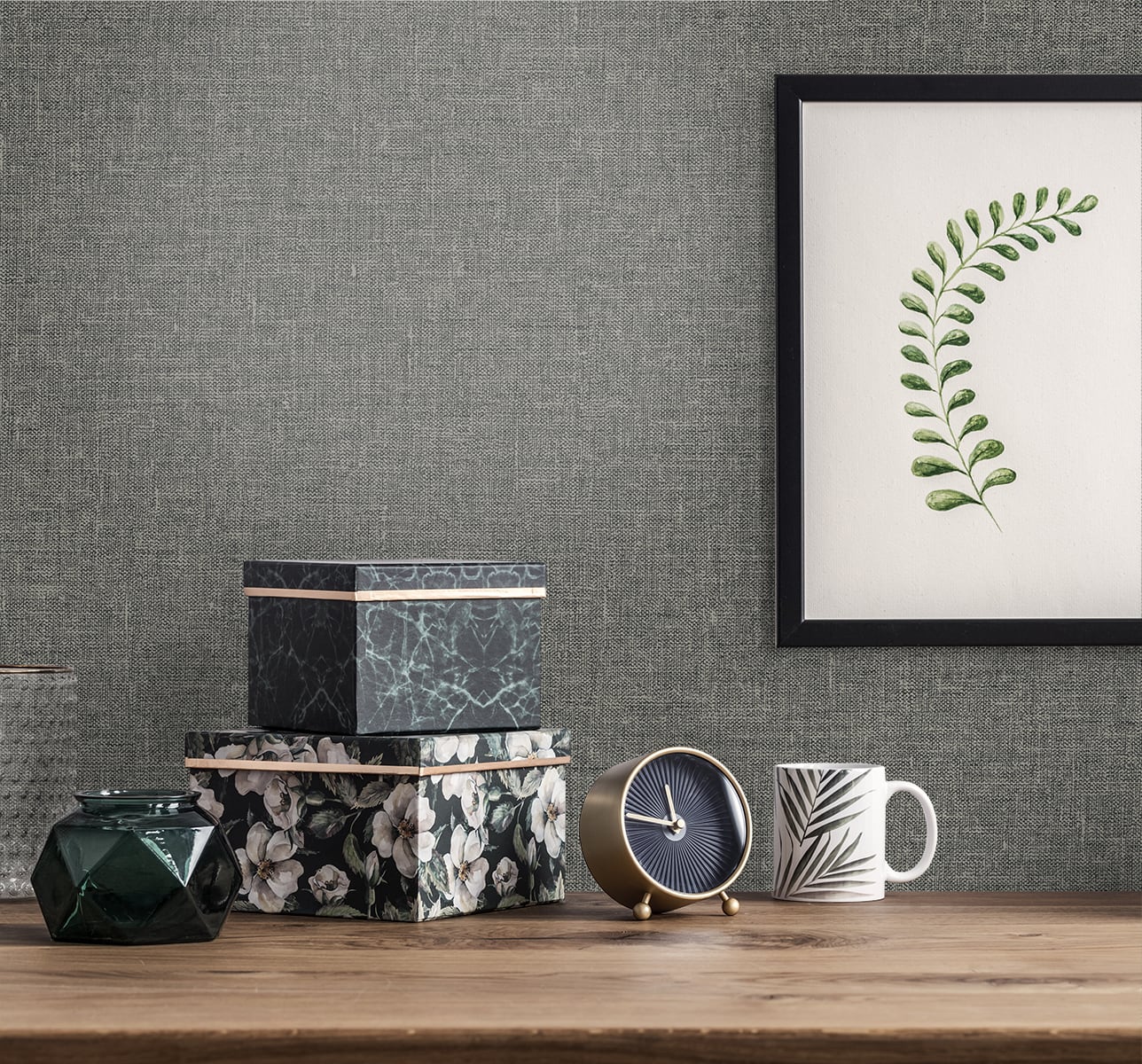 Wooden tabletop with floral and marble boxes, green faceted vase, blue-and-gold clock, striped mug, framed fern print on gray wall.