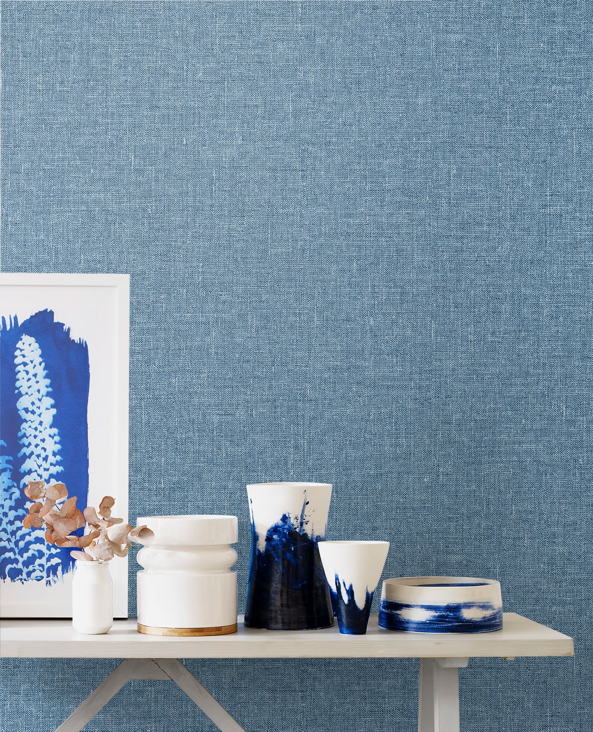 Blue and white linen textile on a light wood table, with framed blue artwork in the background.