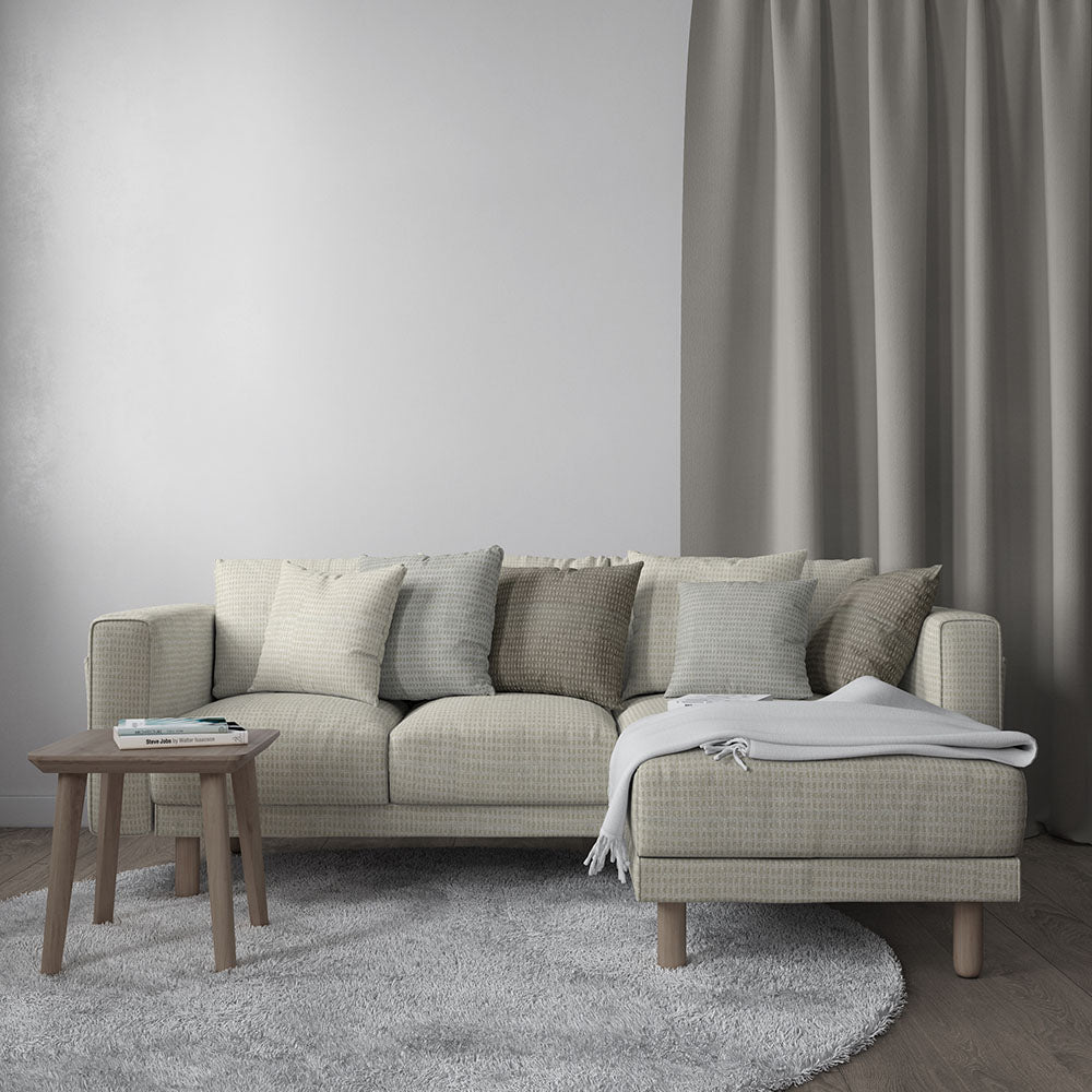 Beige fabric sectional with chaise and wooden legs, neutral cushions, gray rug, round wooden side table, and gray curtains.