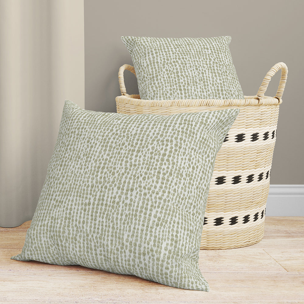 Two sage green solid fabric cushions placed beside a natural woven straw basket with black stripe accents.