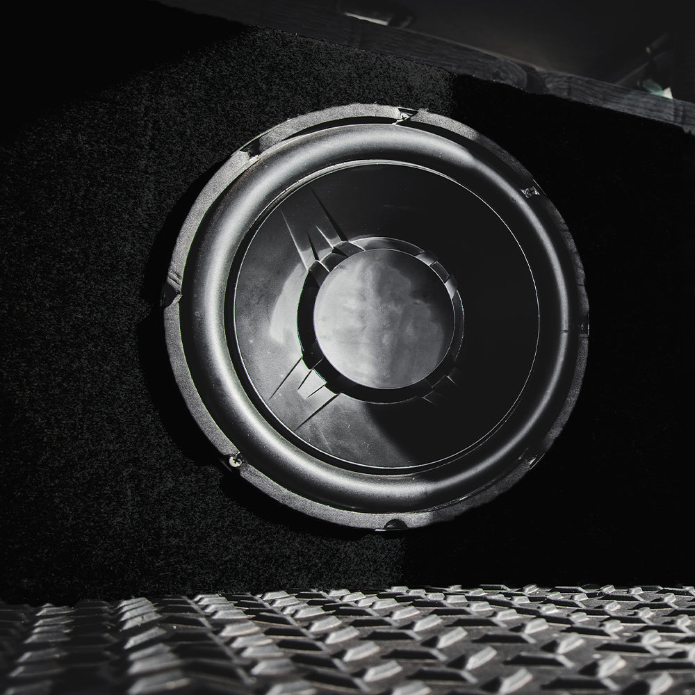 Black speaker with fabric front, circular driver with rubber surround, diamond-plate metal floor visible in the foreground.