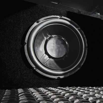 Black speaker with fabric front, circular driver with rubber surround, diamond-plate metal floor visible in the foreground.