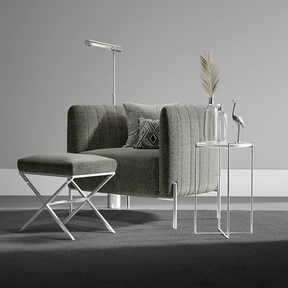 Grey fossil solid fabric upholstery on a tufted armchair with a matching ottoman and chrome legs; glass side tables, floor lamp, vase with palm leaf, crane figurine.