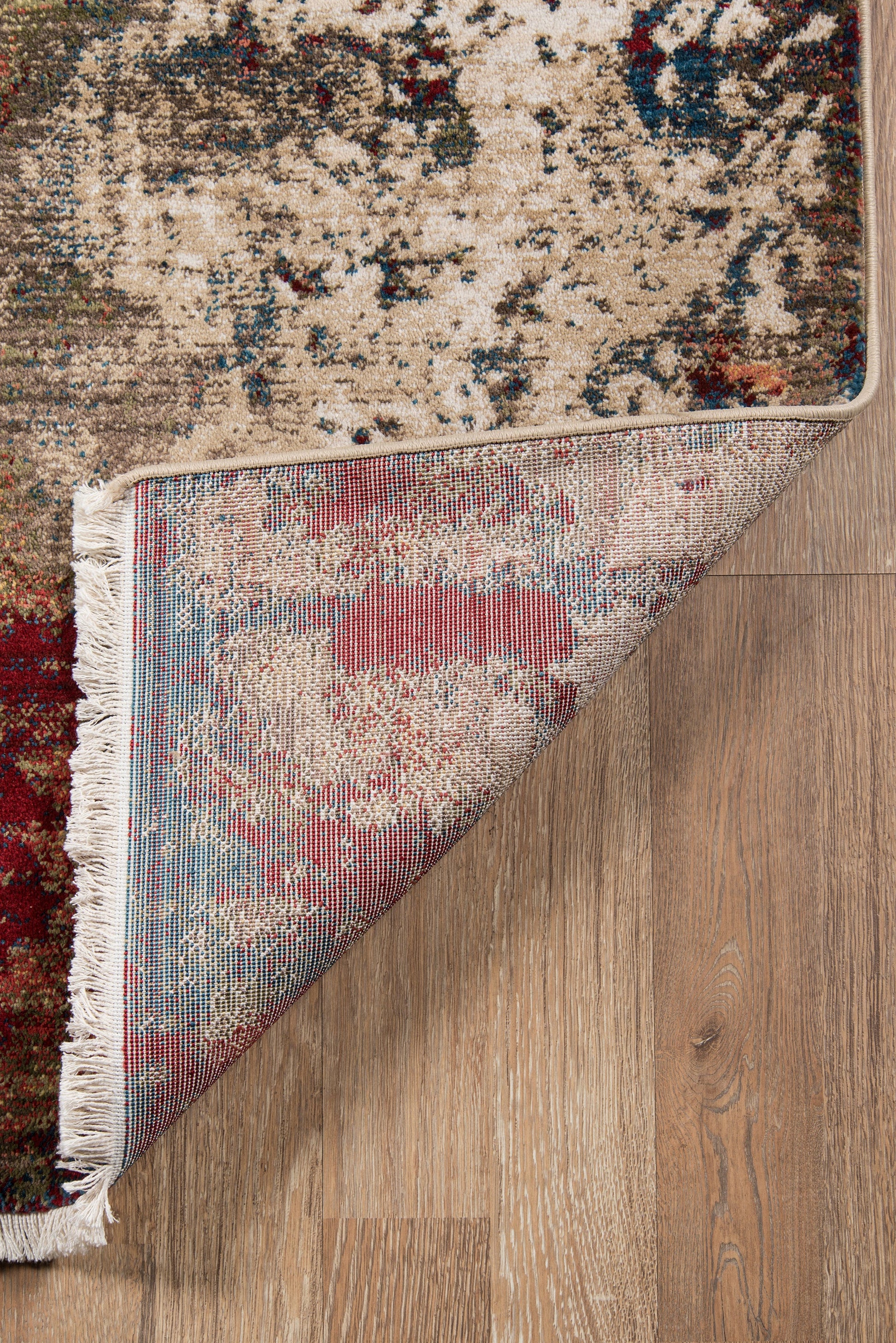 Beige oriental rug with red & blue accents, fringed edge, folded corner reveals patterned underside on wood floor.