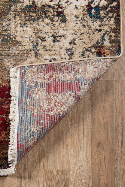 Beige patterned rug with red, blue and cream accents; folded corner shows woven underside and white fringe on wood floor.