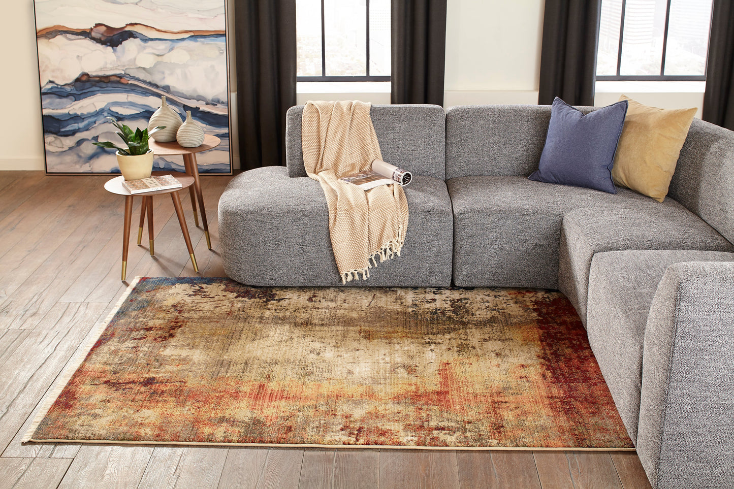 Warm multicolor distressed rug (3' x 5') in a modern living room, with a gray fabric modular sectional, beige throw, and blue/yellow cushions.