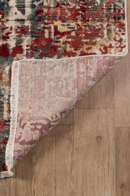Colorful abstract rug with burgundy, cream, blue and gold tones; folded corner, fringe edging on wood floor.