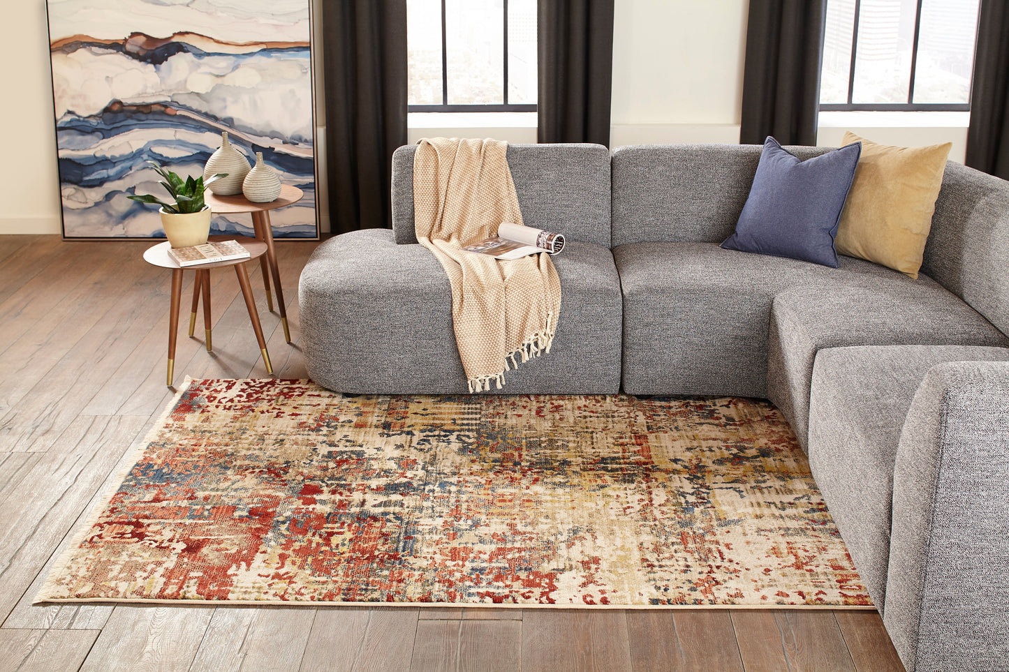 Grey modular sofa with beige knit throw and blue/tan cushions on a colorful distressed rug; round wood side tables with plants.