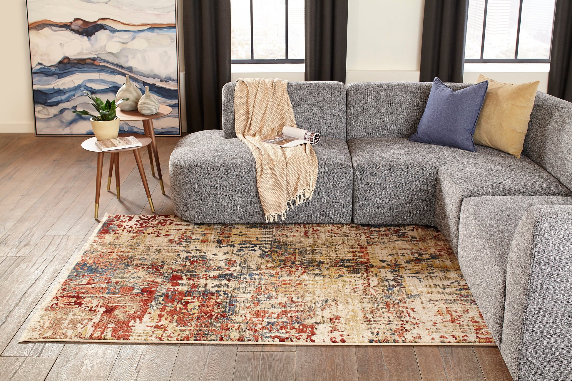 Gray sectional sofa with blue and tan cushions and a beige throw; vintage multicolor rug runner, nesting tables, plant, and abstract wall art.