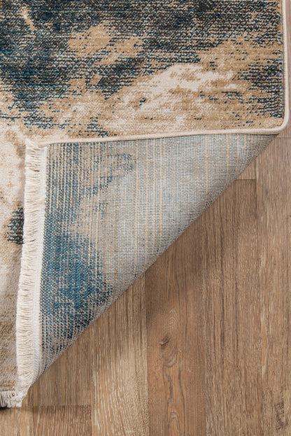 Blue & beige abstract runner rug with fringe along edge, folded corner reveals underside on wood floor.