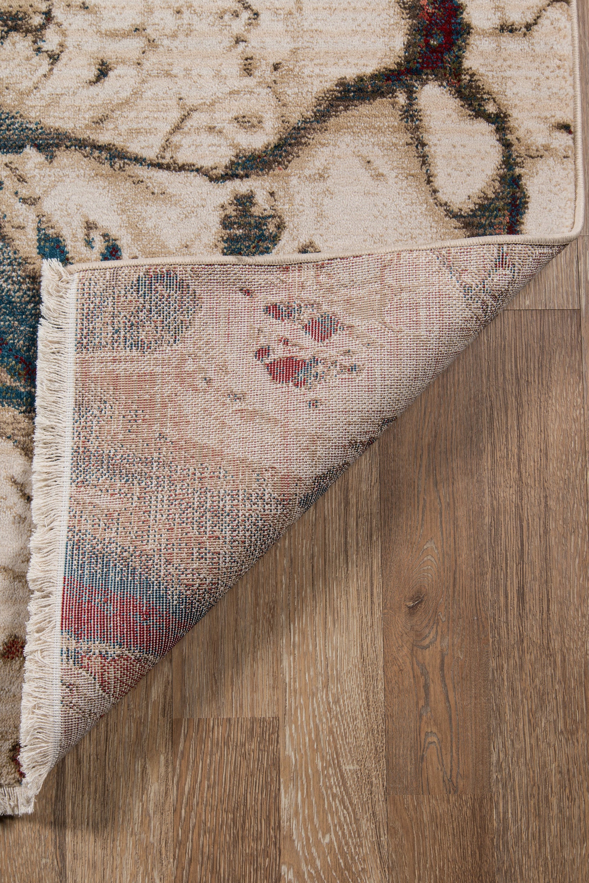Beige oriental rug with blue and red accents, fringed border, folded corner shows underside on wooden floor.