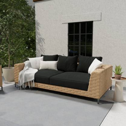 Outdoor rattan sofa with black cushions, white throw pillows, beige wicker frame on a gray patio with potted plants.