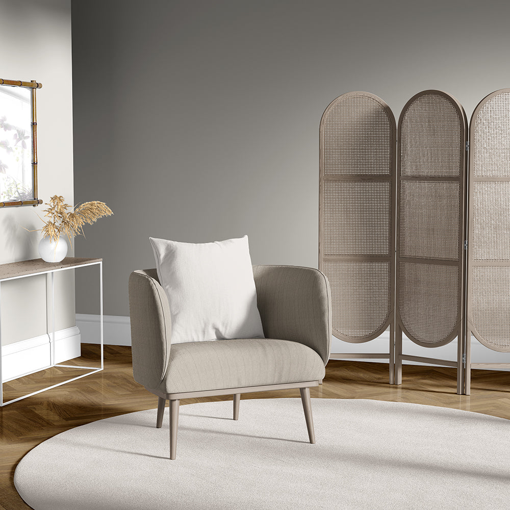 Beige upholstered armchair with a white cushion on a round cream rug; three-panel woven rattan room divider behind.