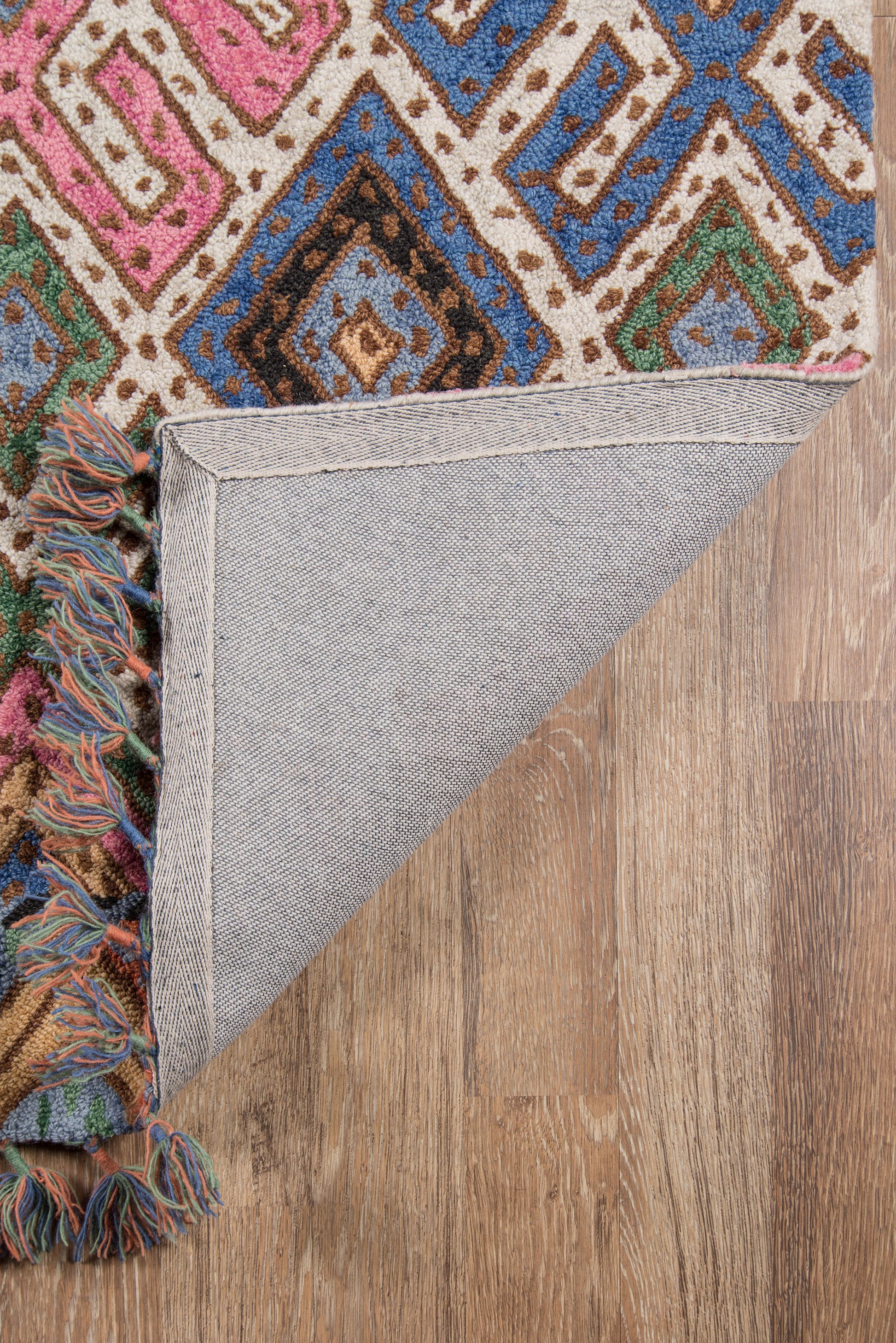 Multicolor geometric rug with diamond motifs; fringed edge, folded corner reveals gray backing on wood floor.