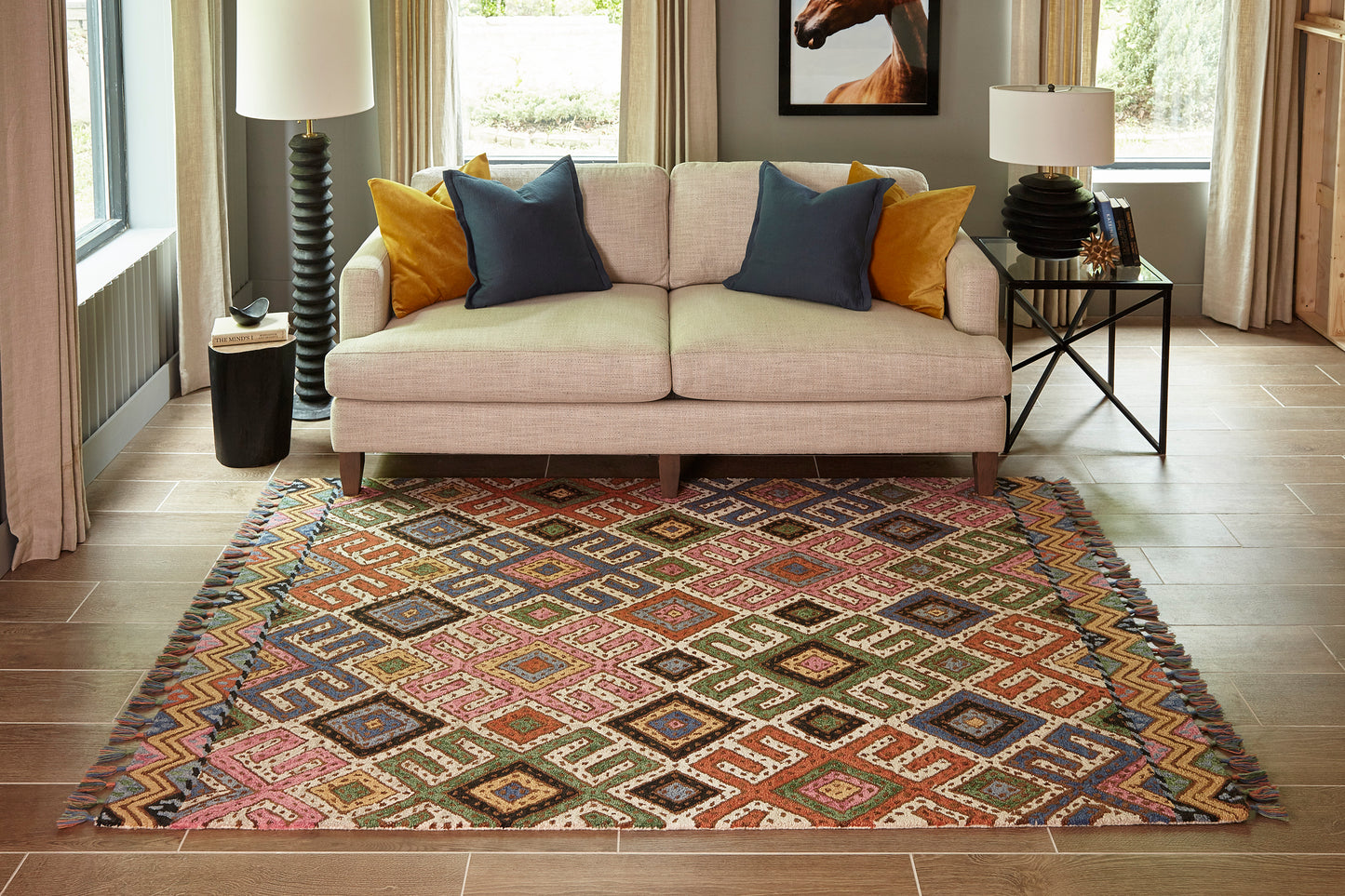 Multicolored geometric diamond rug with fringed edges under a beige sofa, blue and orange pillows, wood floor.