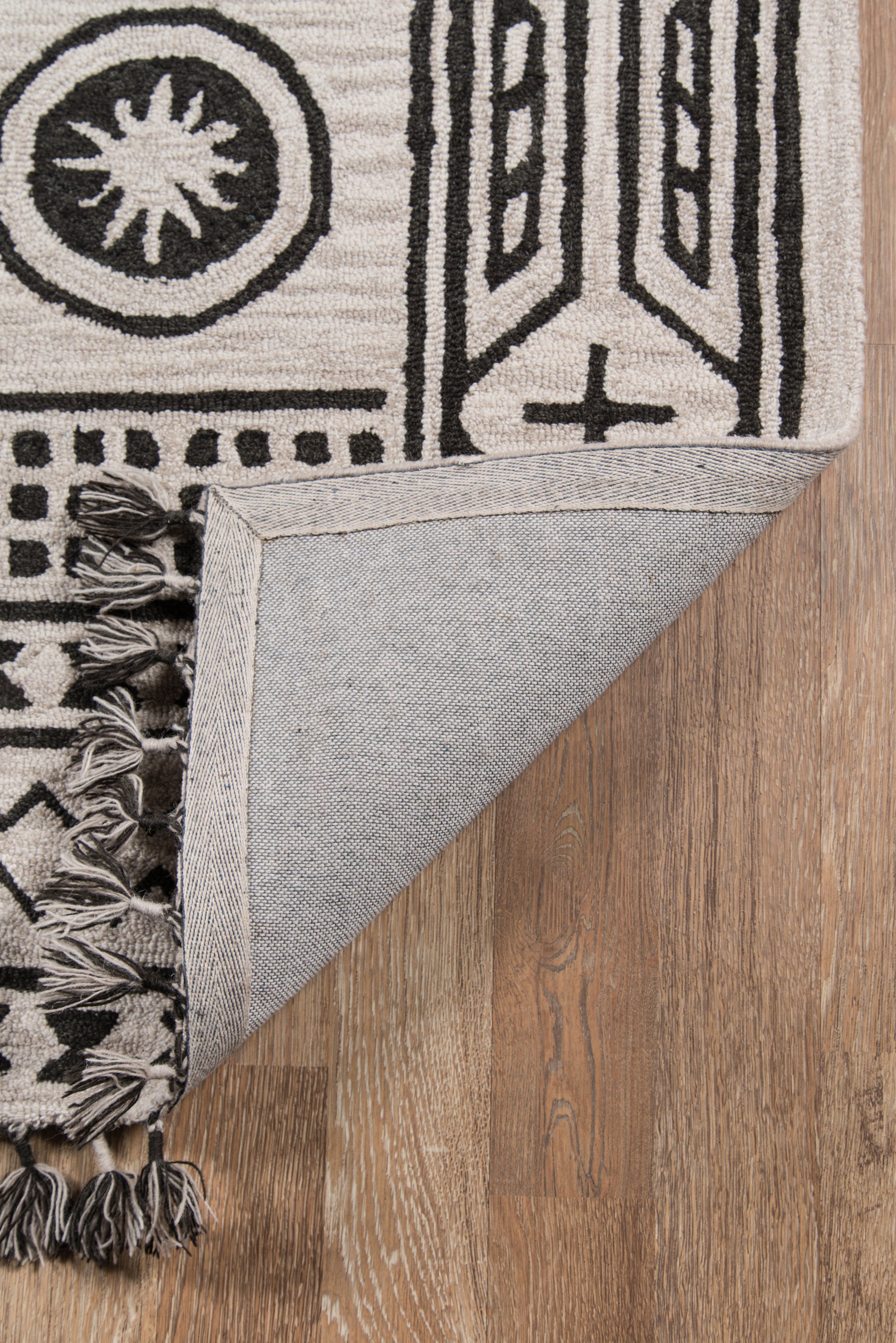 Cream and black geometric rug with fringe tassels; folded corner shows light denim-backed underside on wood floor.