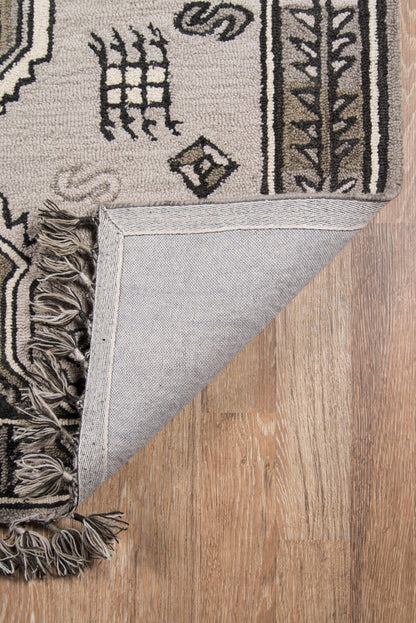 Reversible gray-beige geometric rug with black tribal motifs, fringe tassels, shown half-folded on wood floor.