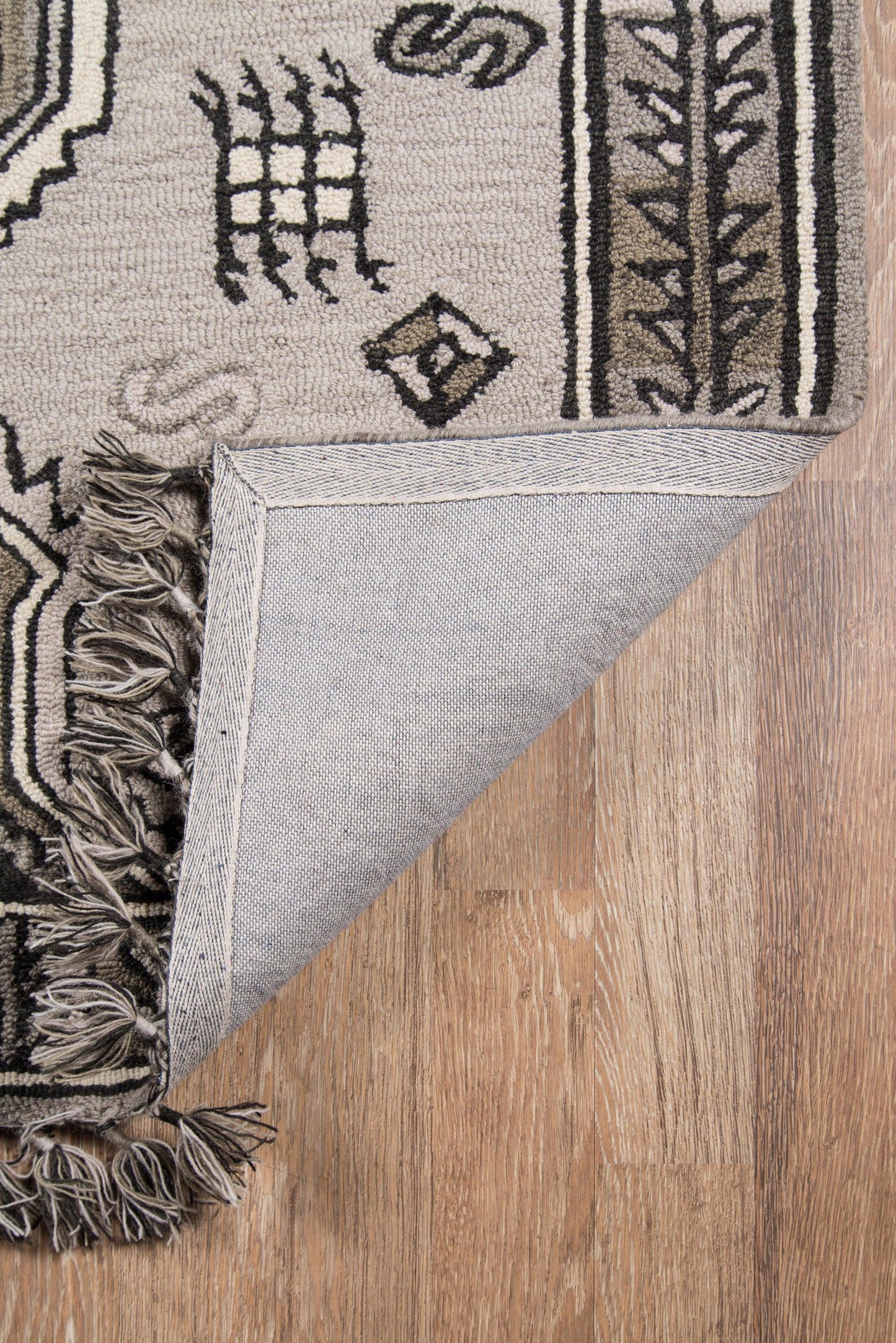 Woven beige and black geometric area rug with fringe trim, folded corner showing light gray underside on wooden floor