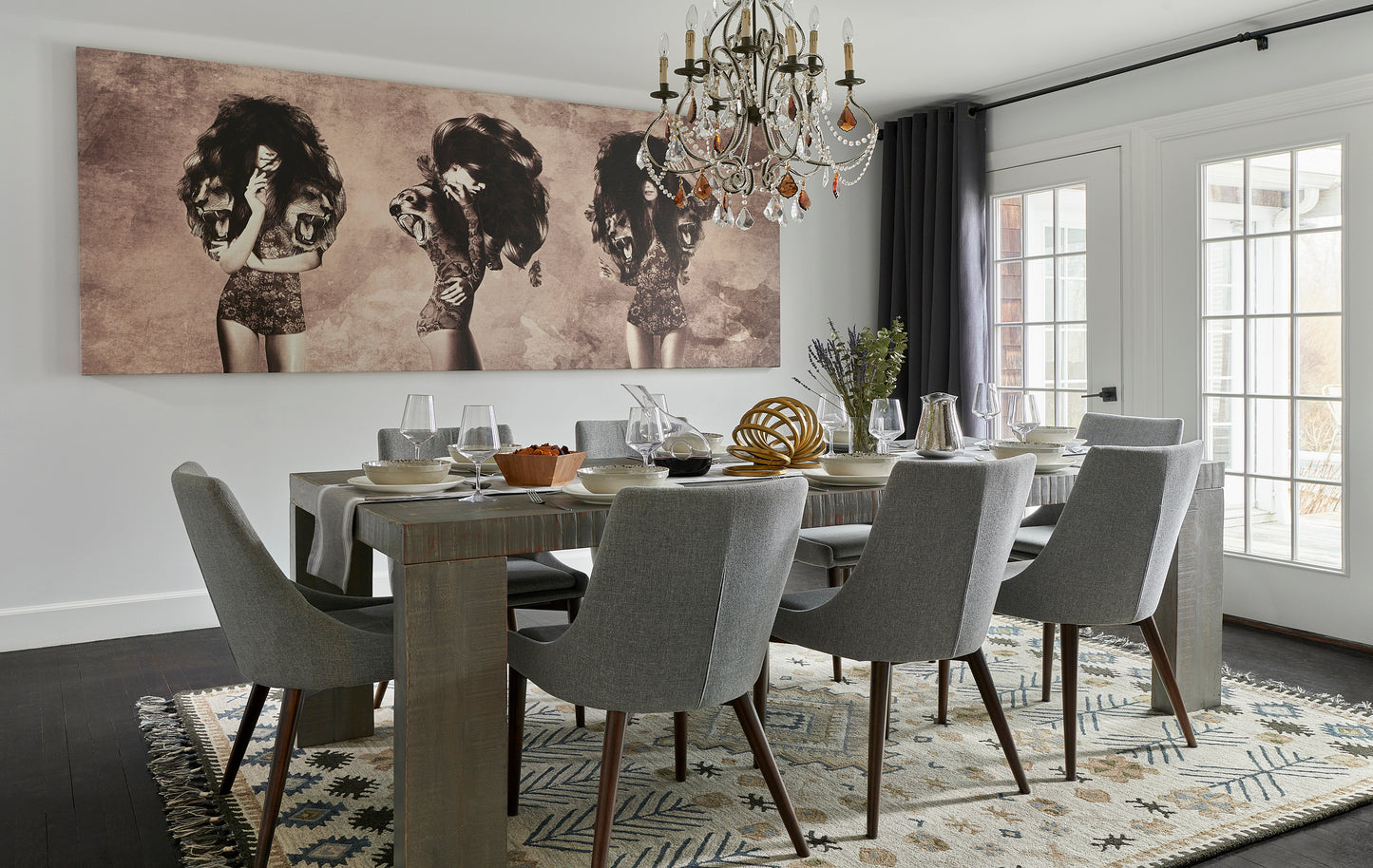 Rustic wooden dining table with gray upholstered chairs, crystal chandelier, beige-blue geometric rug, sepia artwork, dinner setting.