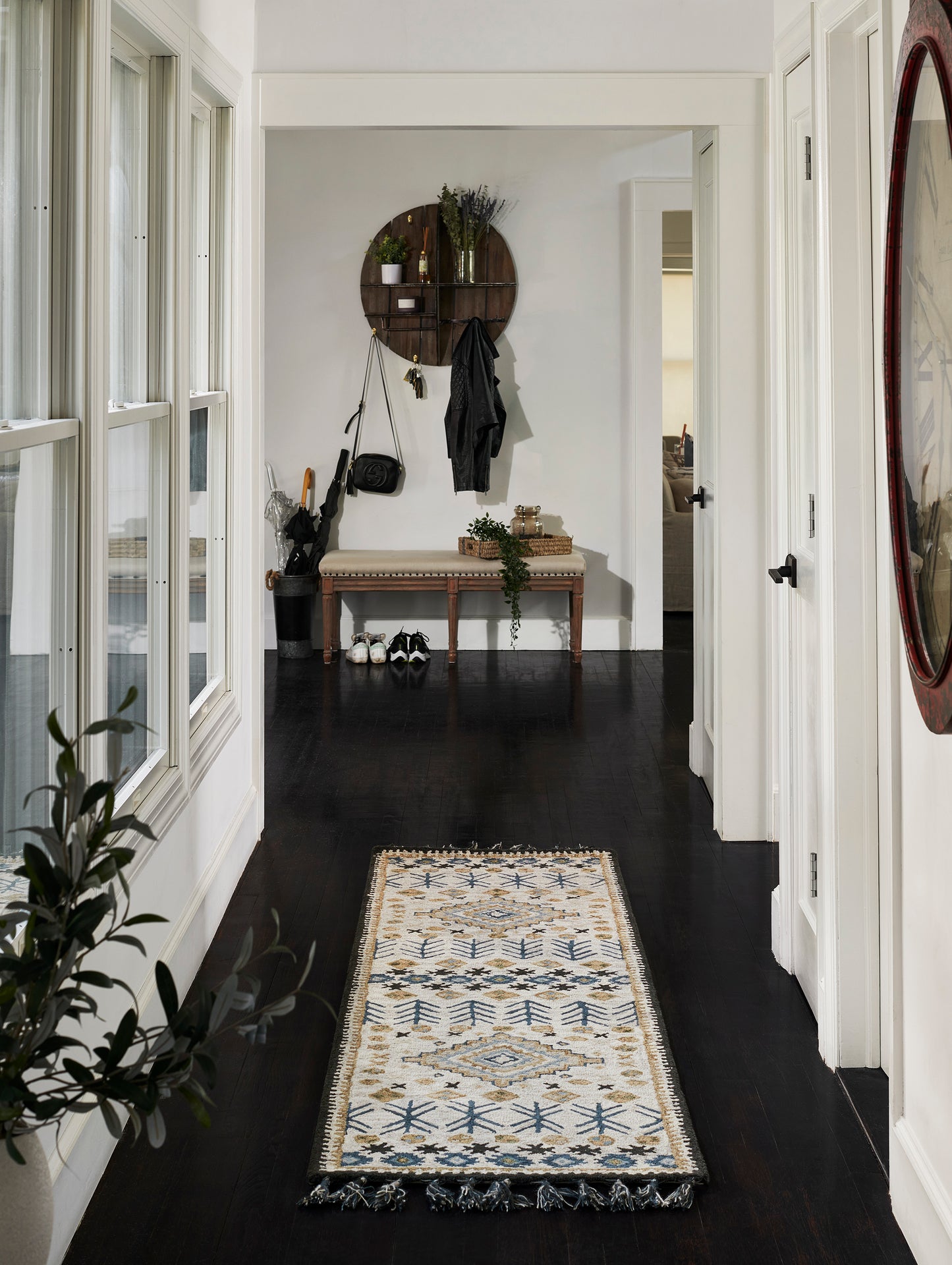 Entryway with dark wood floor, ivory geometric rug, white walls; round wooden wall shelf, coat hooks, bench, plants.
