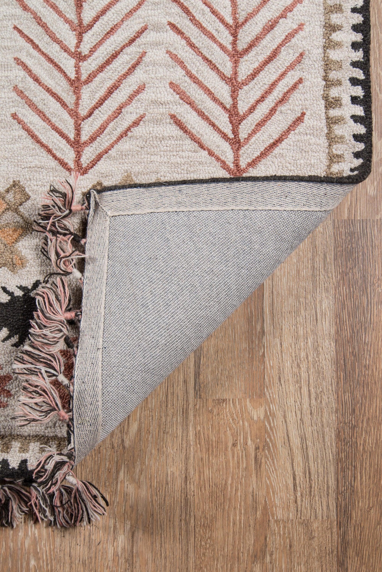 Beige rug with red tree/fern motifs, black border edge, pink tassels; folded corner shows gray underside on wood floor.