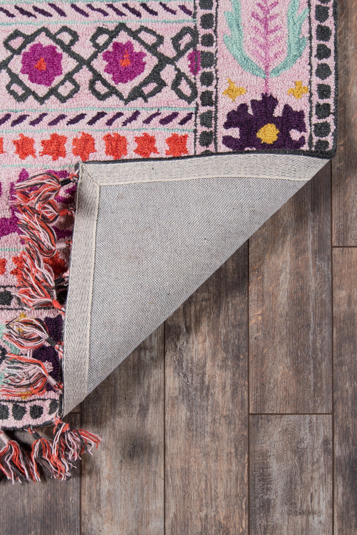 Pink patterned rug with floral motifs in purple and orange; folded corner shows gray woven back and fringe on wood floor.