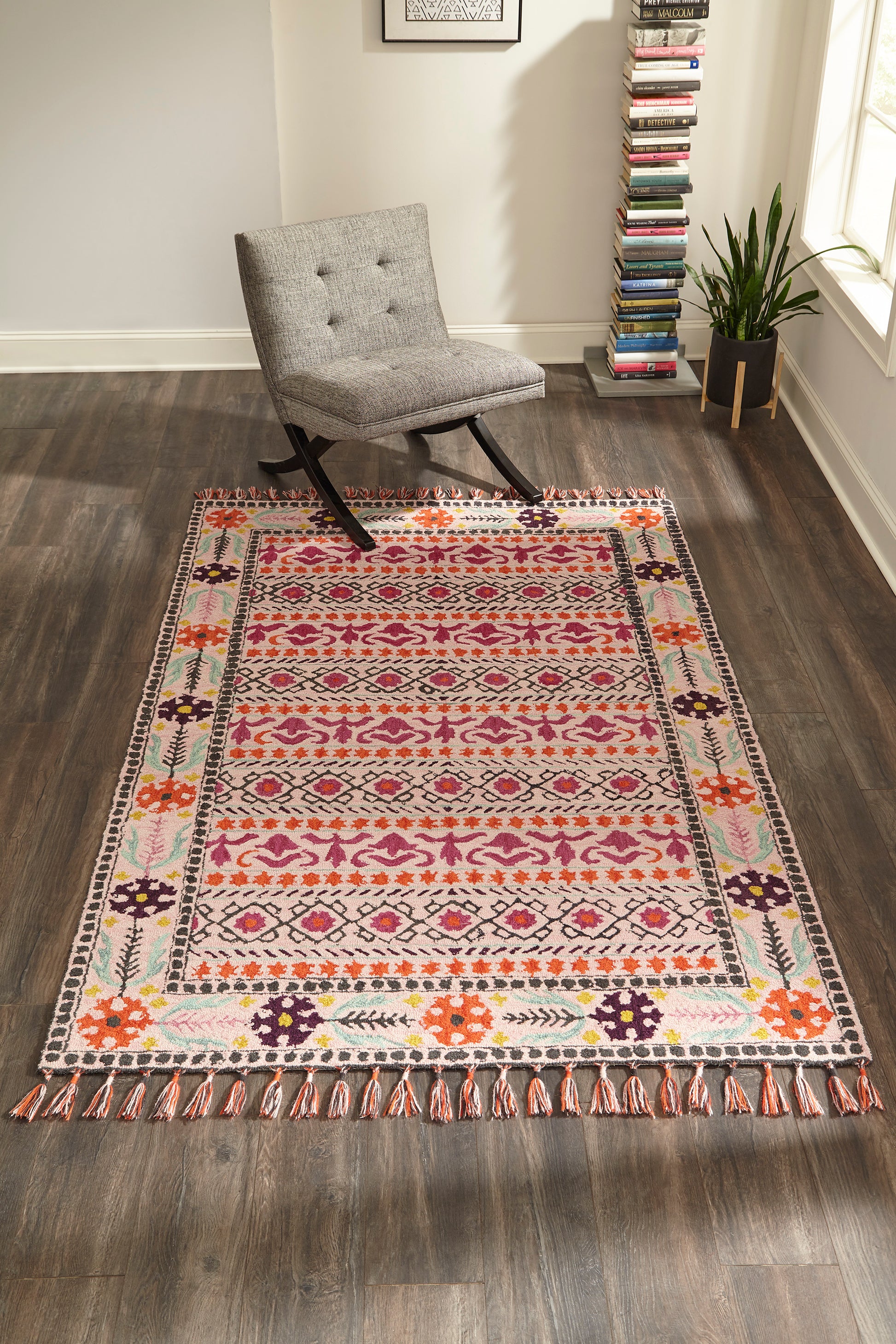 Boho patterned area rug with pink, orange, purple and mint motifs, fringed edges in a living room beside grey tufted chair.