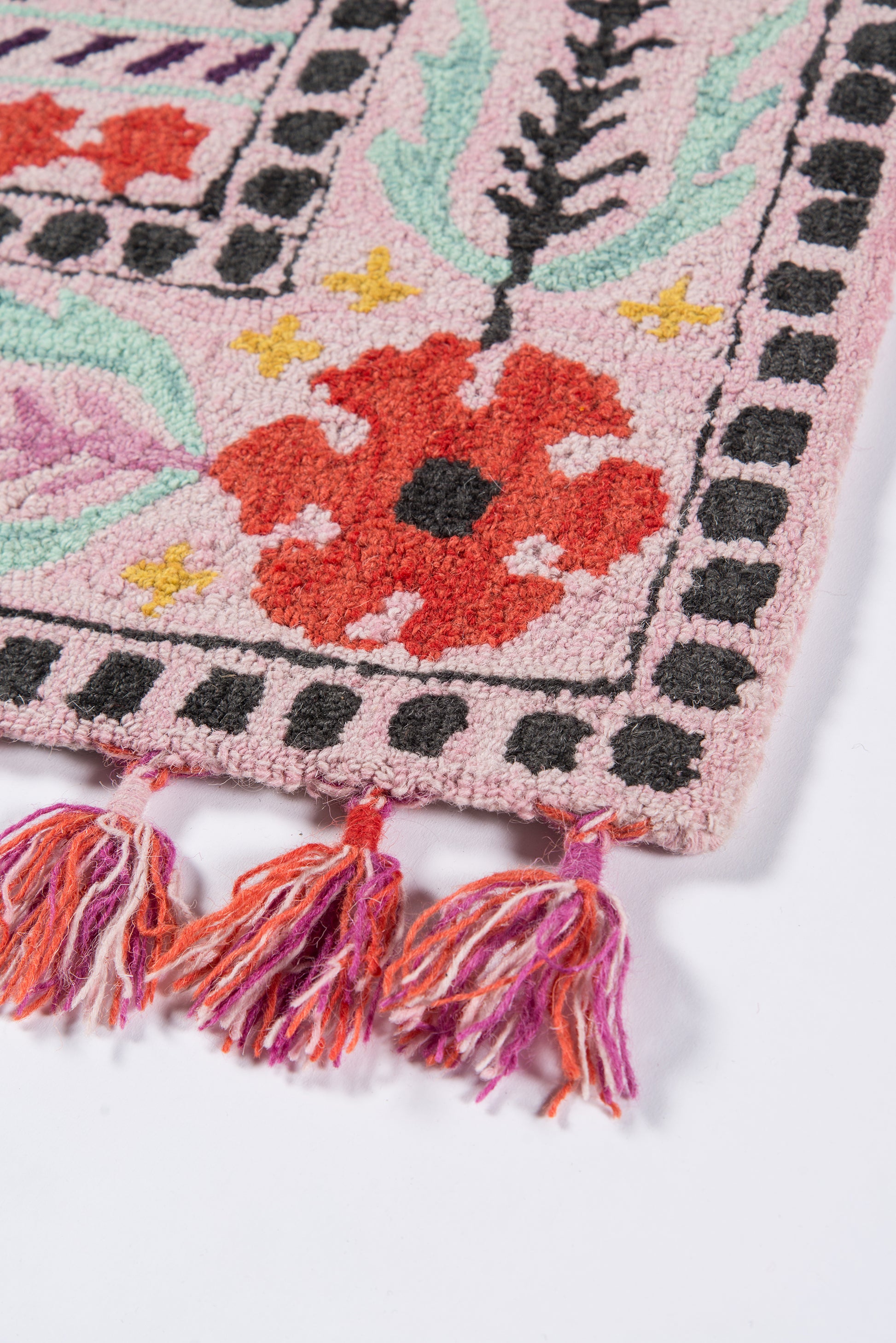 Pink floral woven rug with red flower motif, green leaves, black dotted border, and multicolor tassel fringe.