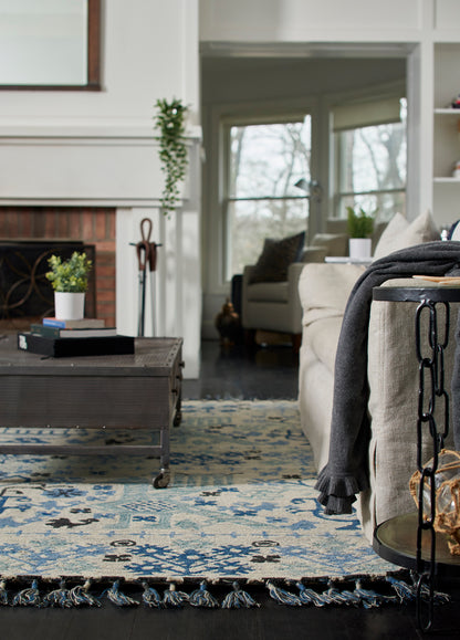 Blue and ivory patterned area rug with fringe in a living room with a dark coffee table and beige sofa with a gray throw.