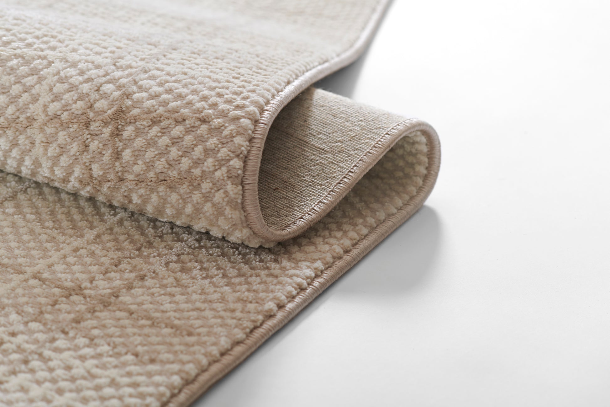 Beige textured loop-pile rug with bound edges, rolled to reveal its underside; soft, neutral rug.