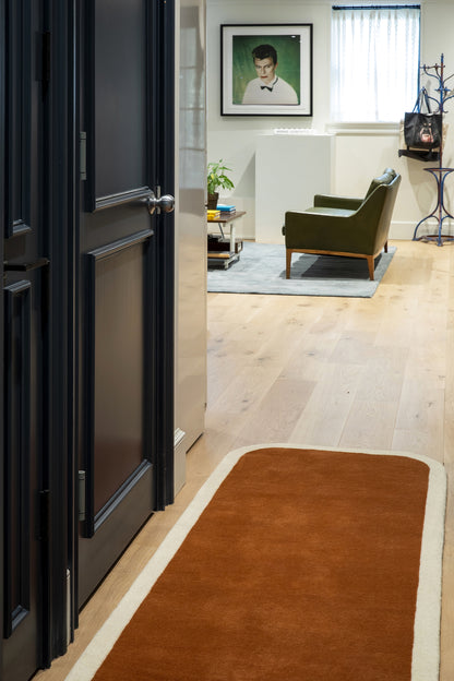 Rust-colored runner rug with cream border along a wooden hallway, opening to a living area with green armchair and framed portrait.
