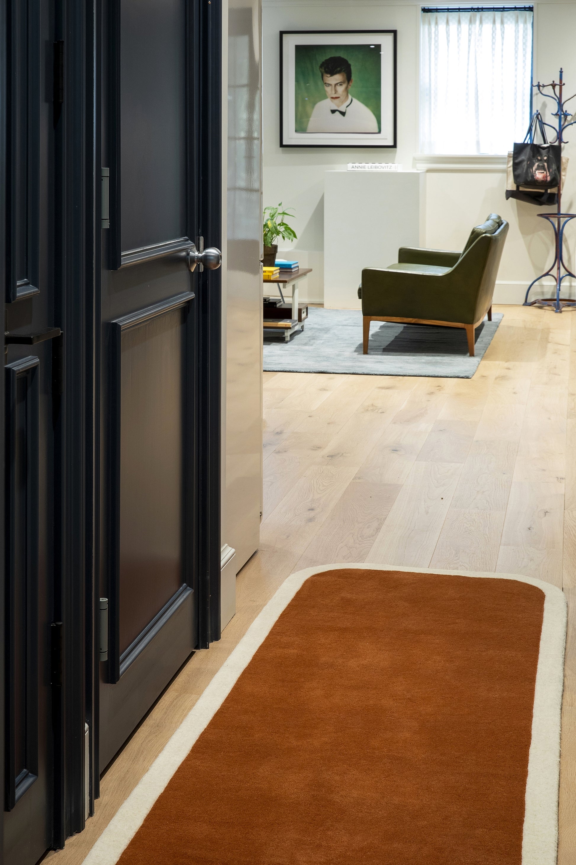 Orange rug runner with cream border on lightwood floor in a living room with green armchair and framed portrait near window.