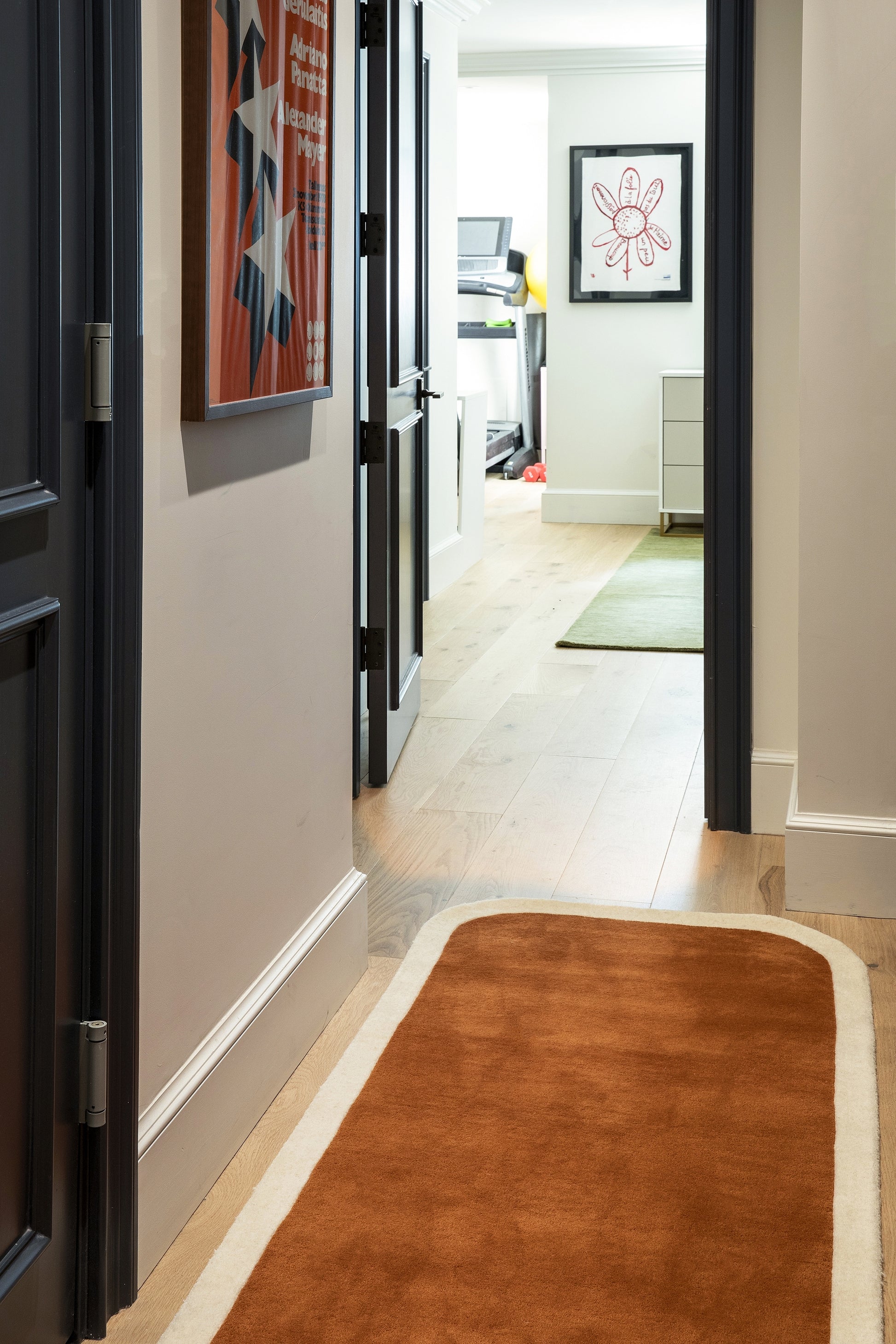 Hallway with light wood floor, dark blue doors, burnt-orange rug runner with beige border, art, leading to a bright room.