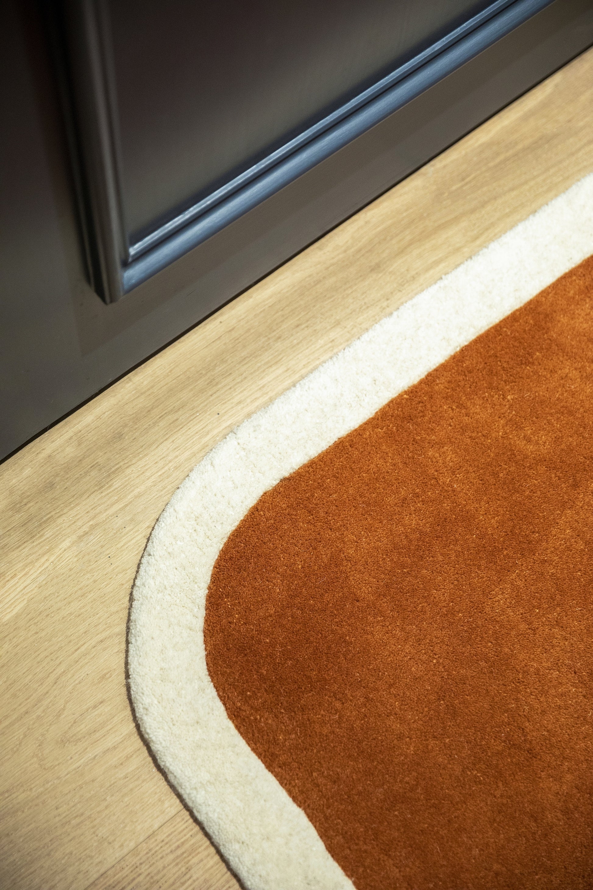 Close-up of rust-orange rug with a cream curved border on light wood floor beside a dark cabinet.