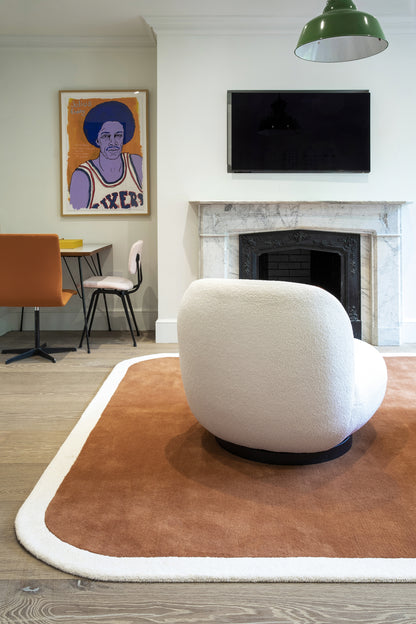 White boucle armchair on a brown rug with white edge; modern living room with marble fireplace, TV, green pendant, art.