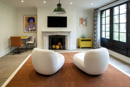 Bright living room with two white curved chairs on a brown rug, stone fireplace with TV, yellow sideboard, windows.