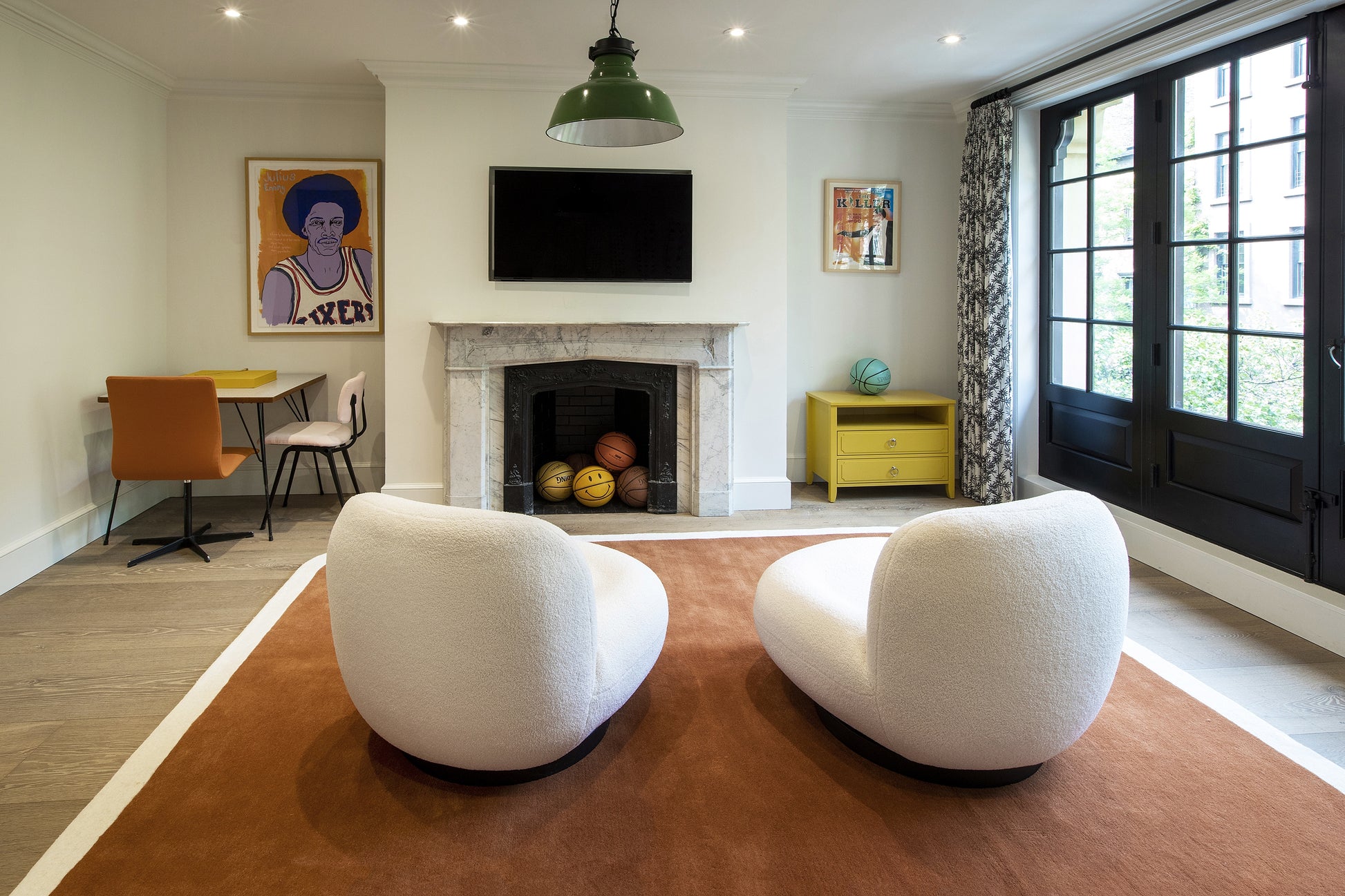 Modern living room with white boucle chairs on a rust rug, marble fireplace, yellow side table, and black-framed windows.