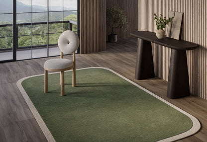 Green rectangular rug with beige border on wood floor; minimalist chair and console table in a sunlit modern living space.