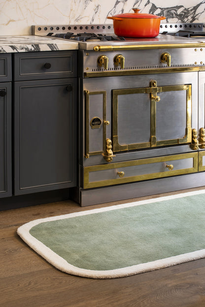 Vintage-style grey kitchen stove with brass trim, red enameled pot on top, dark cabinets, marble backsplash, and green rug.