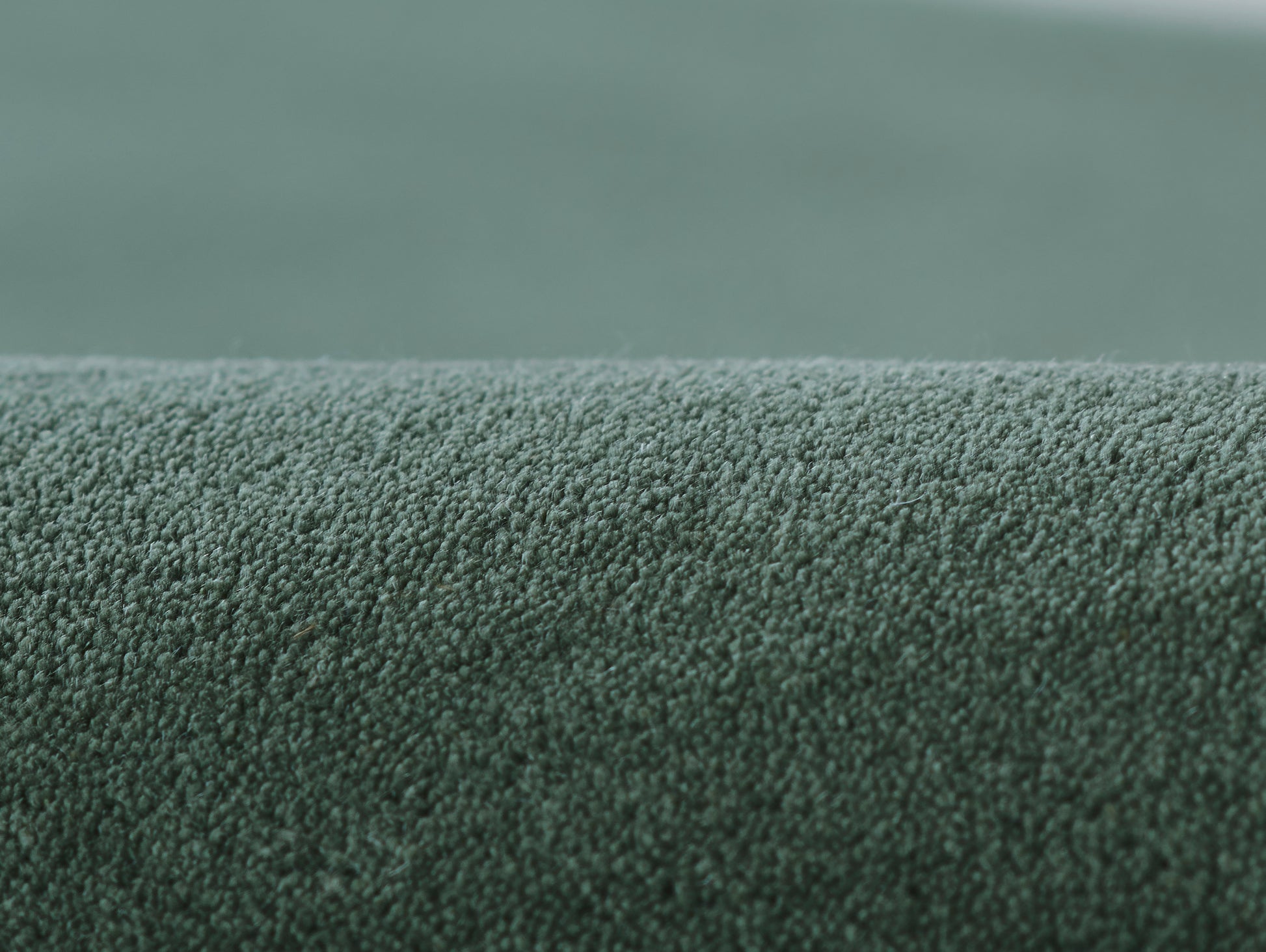 Green loop-pile rug fabric, close-up of dense textured surface with rolled edge.