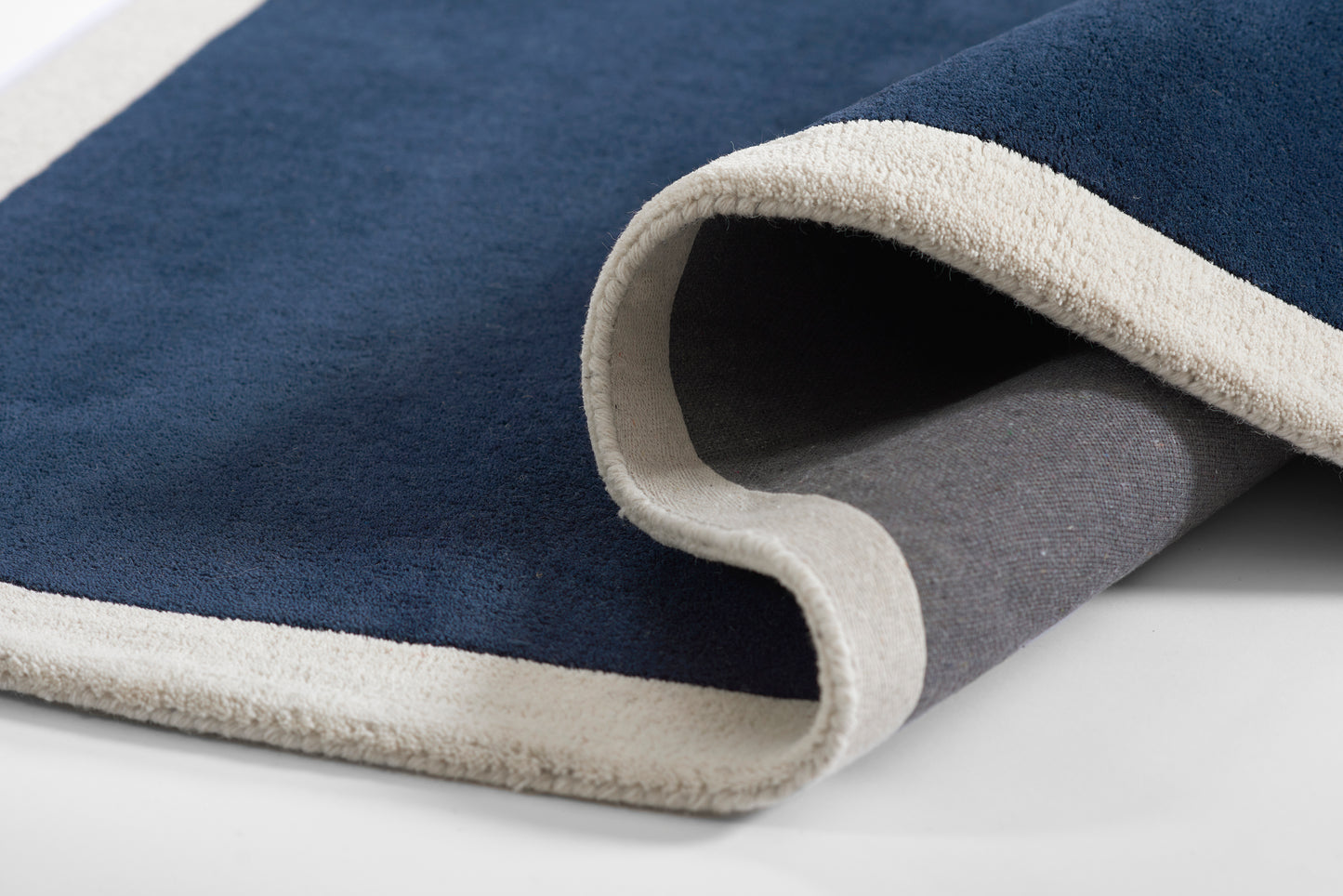 Navy blue rug with beige trim and soft plush pile, partly rolled to reveal grey underside.