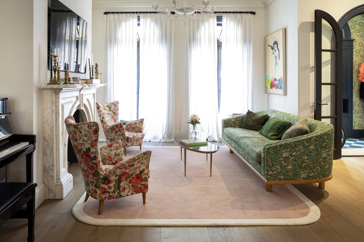 Eclectic vintage living room with green floral sofa, pink armchairs, glass coffee table, fireplace and sheer white curtains.