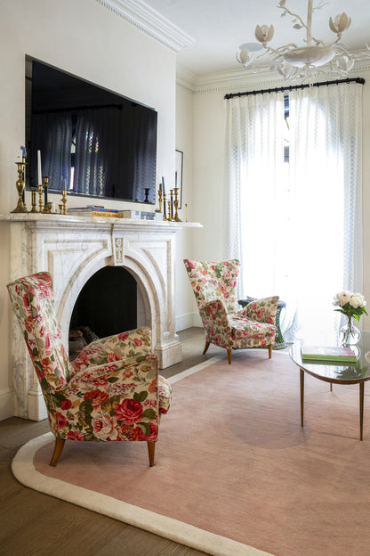 Cozy living room with a white fireplace, pink floral armchairs, glass coffee table, blush rug, TV above mantel, chandelier.