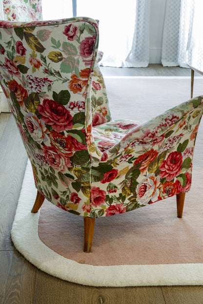 Floral upholstered accent chair with pink roses on light cream fabric, curved back and wooden legs on a beige rug.