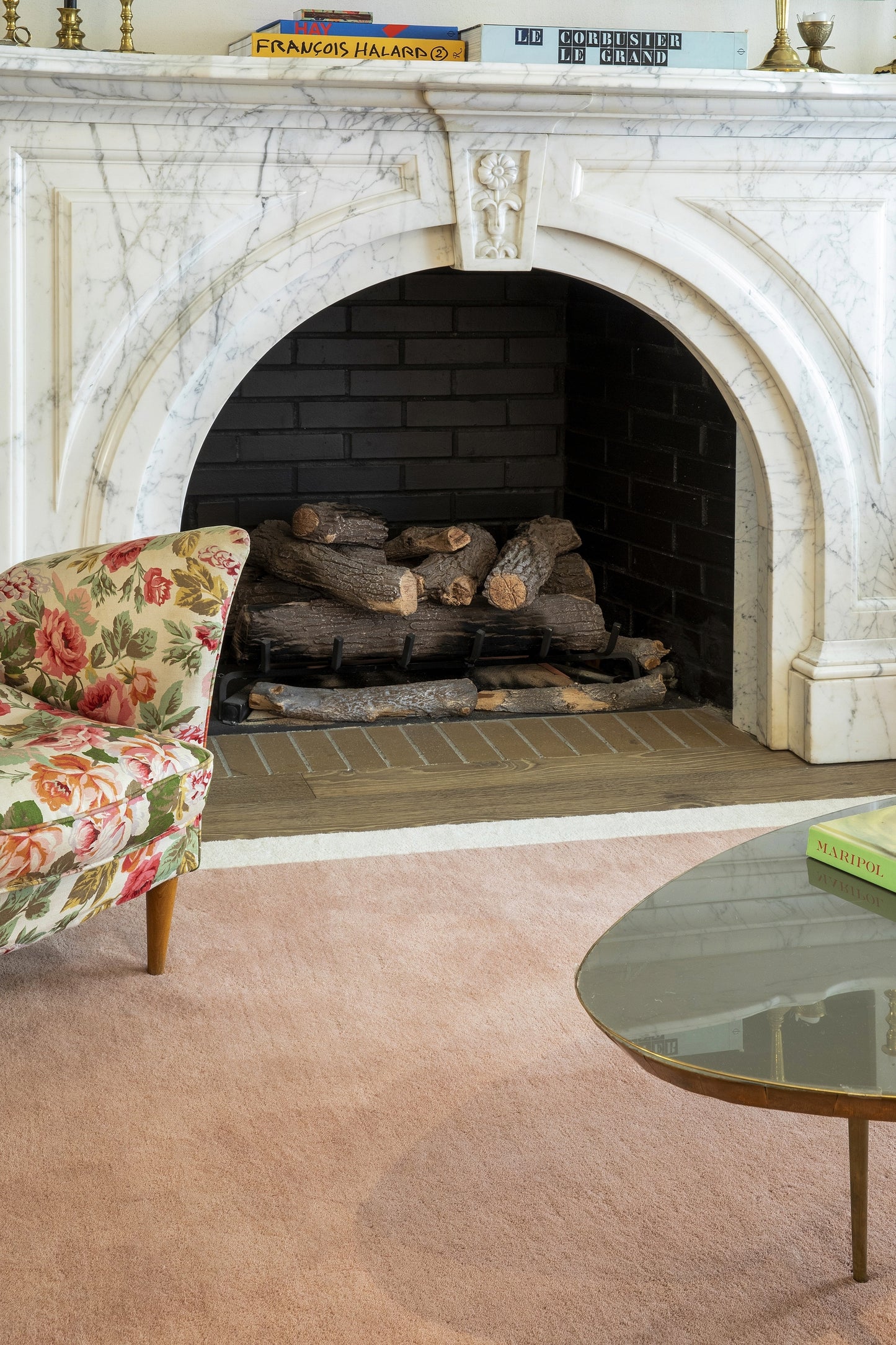 White marble fireplace with stacked logs, floral pink upholstery chair, pink carpet, glass round coffee table, mantel books.
